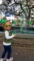 a toddler in st. patricks day attire in front of a green water fountain.