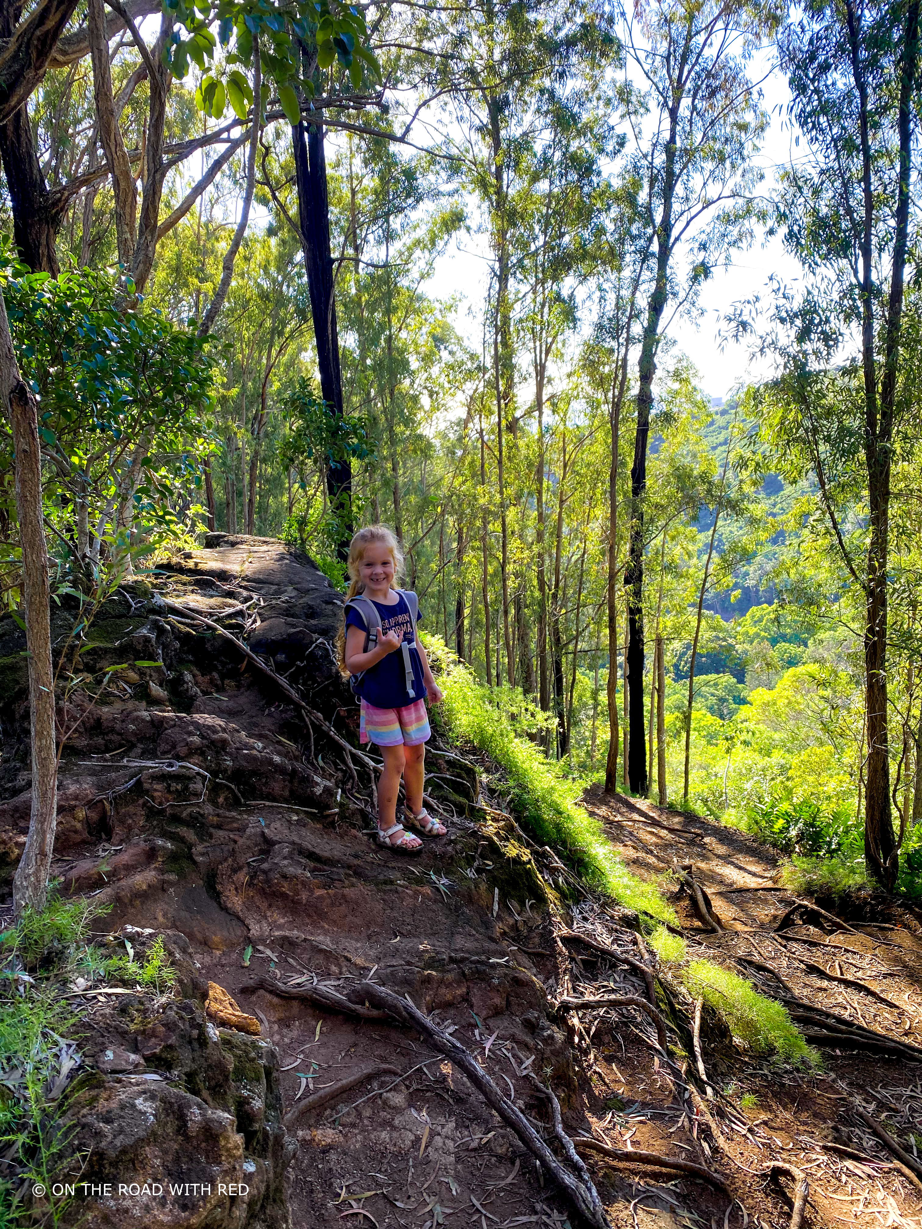 a girl standing on a trail in the hawaiian forest giving a shaka